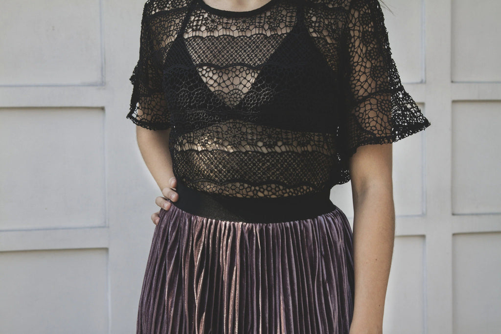 Mujer con blusa negra de encaje transparente y falda plisada violeta frente a pared blanca