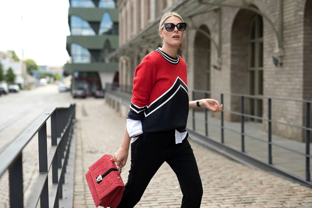 Mujer con suéter rojo y negro, gafas de sol y bolso rojo caminando en calle empedrada urbana