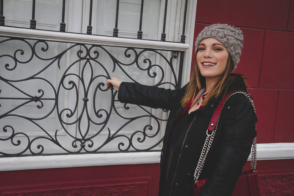 Mujer sonriente con gorro gris y chaqueta negra junto a ventana con reja decorativa de hierro