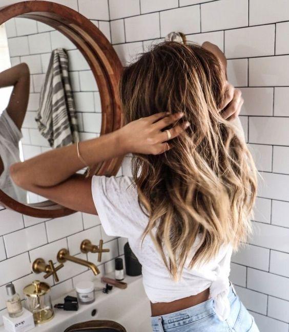 Mujer con cabello rubio ondulado peinándose frente a un espejo redondo en baño moderno con azulejos blancos