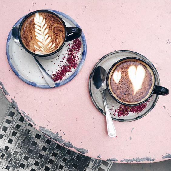 Dos tazas de café con arte latte en platos cerámicos sobre mesa rosa, con cucharas y decoración de flores secas
