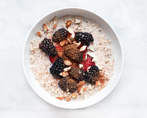 Tazón blanco con avena, moras negras, fresas y almendras picadas sobre fondo blanco