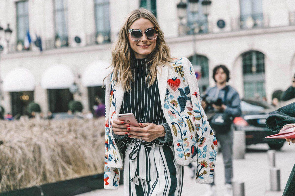 Mujer con gafas de sol grandes y chaqueta blanca bordada con flores, usando teléfono móvil en calle urbana